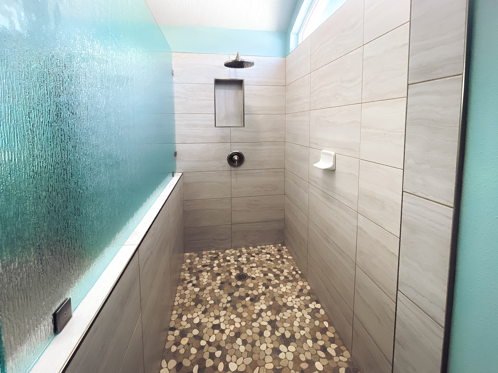 Modern shower with textured glass panels, light gray tiled walls, a rainfall shower head, and pebble stone flooring.