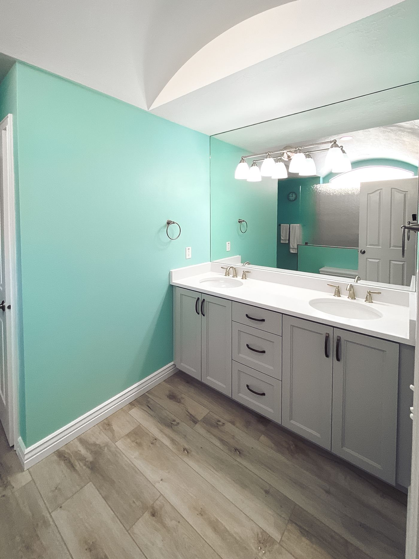 Modern bathroom with a double vanity featuring gray cabinetry, white countertop, and gold faucets, paired with light turquoise walls and wood-look tile flooring.
