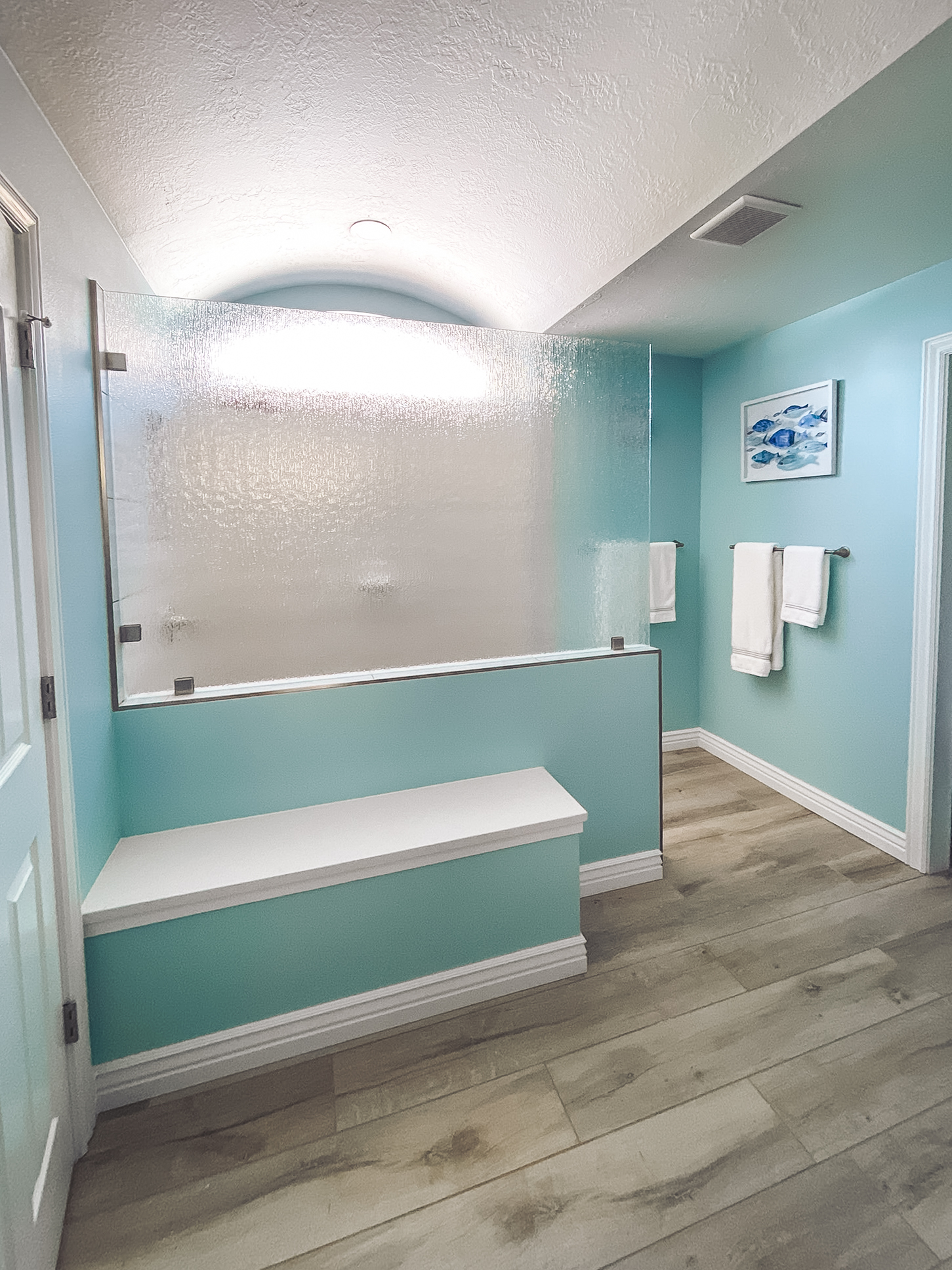 Modern bathroom with a frosted glass shower partition, light turquoise walls, and a white bench, featuring wood-look tile flooring.