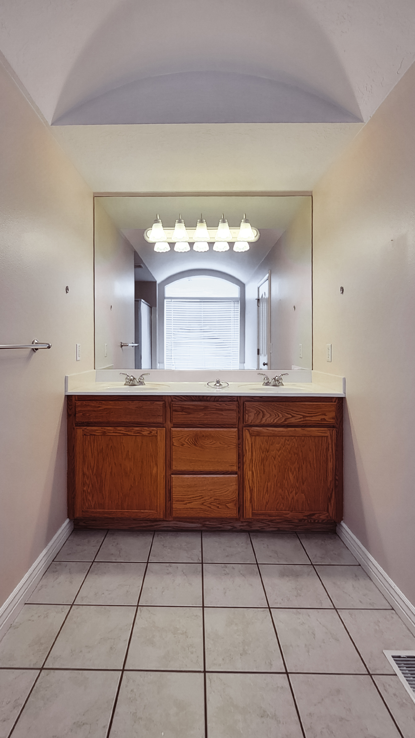 Bathroom with an oak wood vanity, double sinks, and a large mirror, with beige tile flooring and a window letting in natural light.