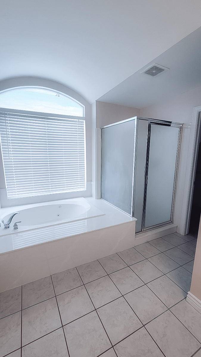 Bathroom with a white bathtub under an arched window, a glass shower stall, and beige tiled flooring.