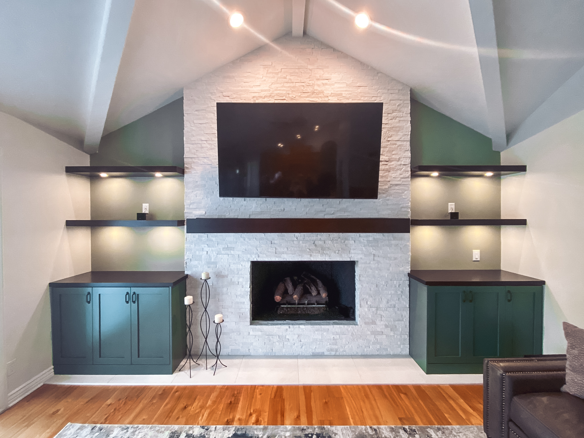 Modern living room with a stone fireplace, sleek dark green cabinetry, floating shelves with lighting, and a mounted TV above the fireplace.