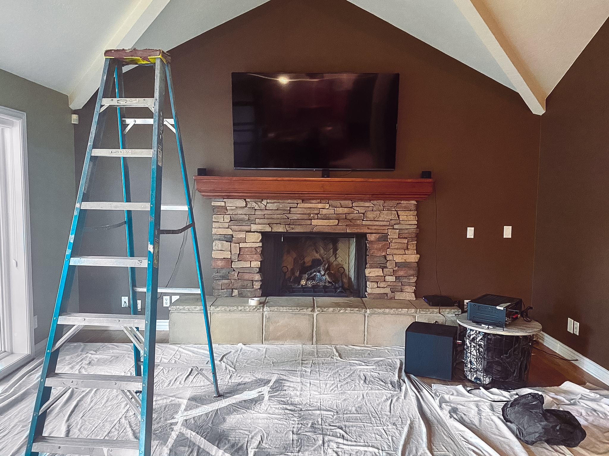 Living room with a stone fireplace, wooden mantle, TV mounted above, and a ladder placed on a drop cloth for renovation work.