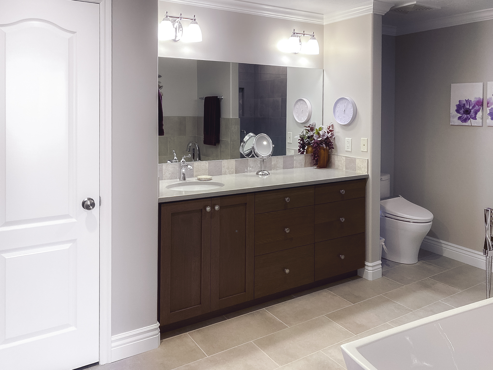 Modern bathroom featuring a sleek vanity with a quartz countertop, large mirror, and contemporary cabinetry, with beige tile flooring and a toilet in the background.