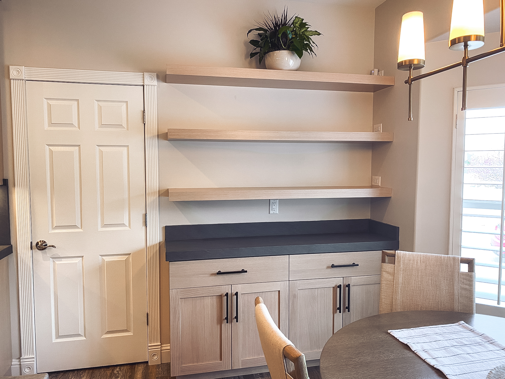 Modern kitchen nook with light wood cabinetry, black hardware, dark stone countertop, and floating shelves.
